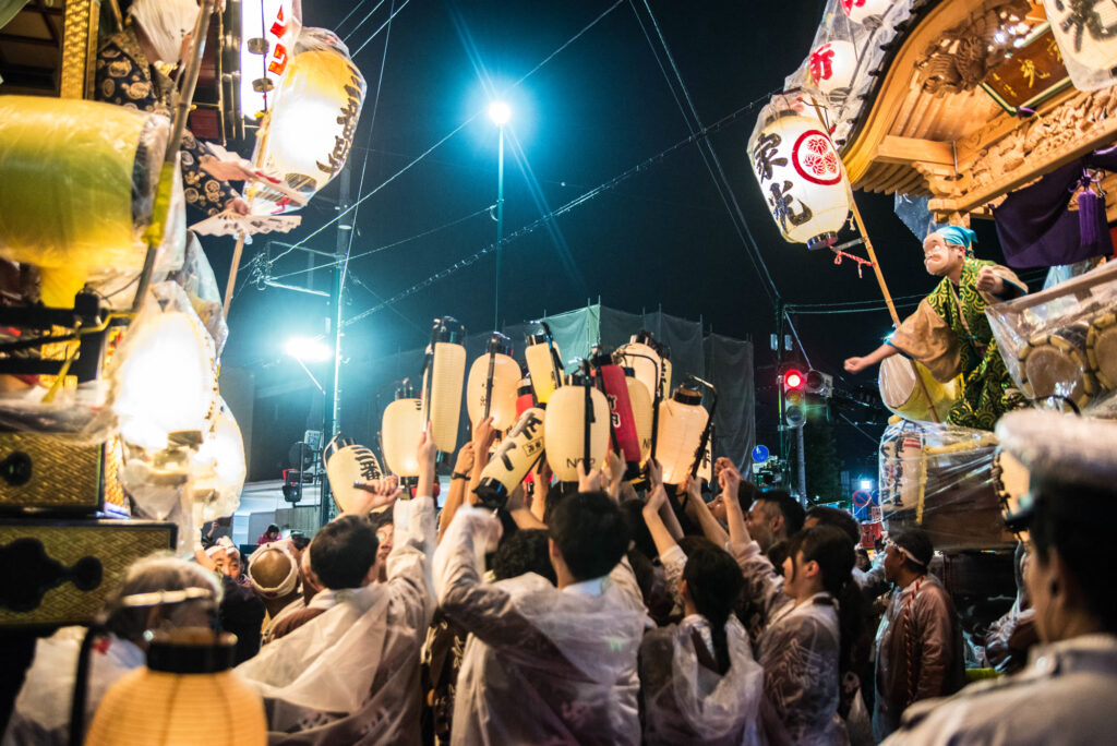 向かい合う2台の山車と提灯を掲げる人たちを、横から低い位置で撮った写真