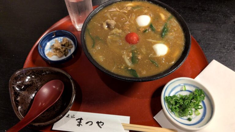 麦匠 まつや（豊橋）の豊橋カレーうどん。黒い器にうずら卵2つとミニトマトがのり、薬味のねぎと漬物がつく